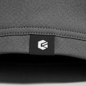 Closeup of a small, custom black and white woven label with a logo attached to the bottom hem of a textured dark grey softshell jacket.