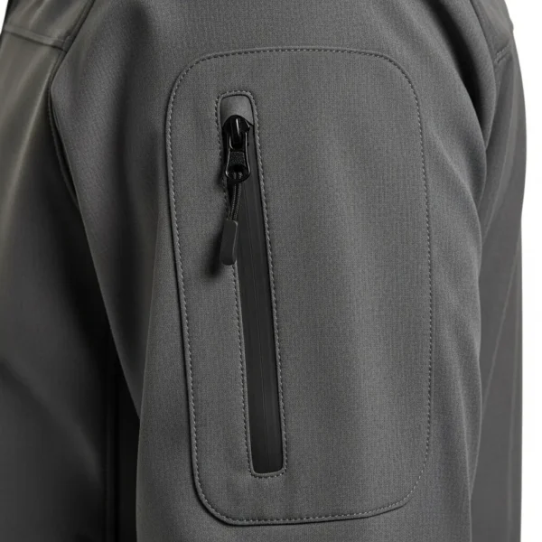 Closeup of a dark grey softshell jacket sleeve showing a functional, vertical zippered arm pocket with a black waterproof seam.